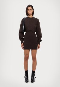 Brun strikket minikjole med lange puffærmer, indsnørret talje og knapdetalje, parret med sorte ankelstøvler og en sort håndtaske.