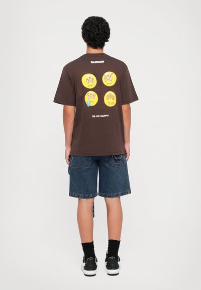 Osoba v hnedom tričku so štyrmi žltými roztrhnutými smajlíkmi a textom "JSEM TAK ŠŤASTNÝ" na chrbte, v kombinácii s denimovými šortkami.