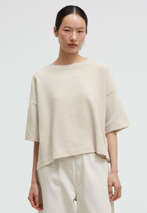 Frau mit dunklen zurückgebundenen Haaren, die ein beiges, locker sitzendes, geripptes Kurzarmoberteil und offwhite, hoch taillierte Hosen vor einem schlichten Hintergrund trägt.