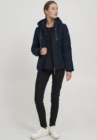Doudoune marine avec une capuche, fermeture éclair frontale et accents de cordon en marron. Portée sur un haut noir et assortie à un pantalon noir et des baskets blanches.