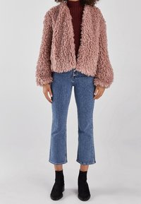 Faux fur-jacka i mjuk rosa, öppen framtill, med stora ärmar. Kombinerad med blå denimflare jeans och svarta ankelstövlar.