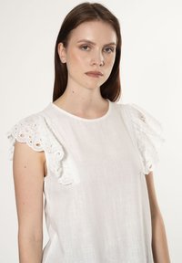 Blusa bianca in tessuto testurizzato, con maniche a balza in pizzo e scollatura rotonda, finitura liscia e senza chiusure visibili.