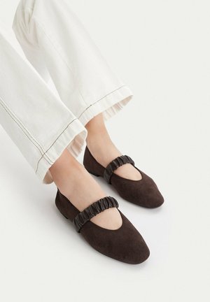 Pies con zapatos planos Mary Jane de ante marrón oscuro con correas elásticas fruncidas y pantalones blancos cortos con costuras visibles.