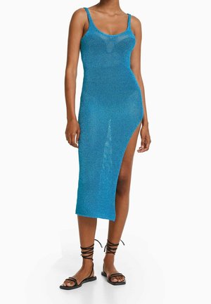 Femme portant une robe en maille bleue sans manches avec une fente haute sur le côté, associée à des sandales plates noires à brides.