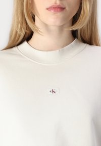 Light beige crewneck sweatshirt with ribbed collar, featuring a small rectangular logo patch in white and burgundy on the left chest.