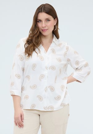 Frau mit langen braunen Haaren, die ein weißes Hemd mit beigen Paisley-Mustern und hellbeige Hose trägt, posiert mit einer Hand in der Hüfte.