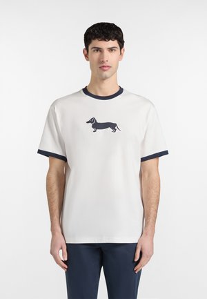 Młody mężczyzna w białej koszulce z granatowym nadrukiem jamnika na piersi oraz granatowymi wykończeniami na rękawach i kołnierzu, stojący na białym tle.