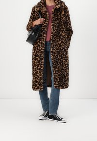 Lange jas met luipaardprint van nepbont, met een ruime kraag en een open voorkant. Draagt het over een bordeauxrode blouse, blauwe spijkerbroek en zwarte sneakers.