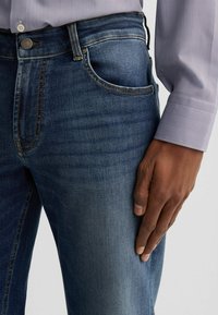 Jeans in denim blu scuro con cuciture a contrasto, una tasca frontale e chiusura con bottone. Una mano poggia sulla coscia.