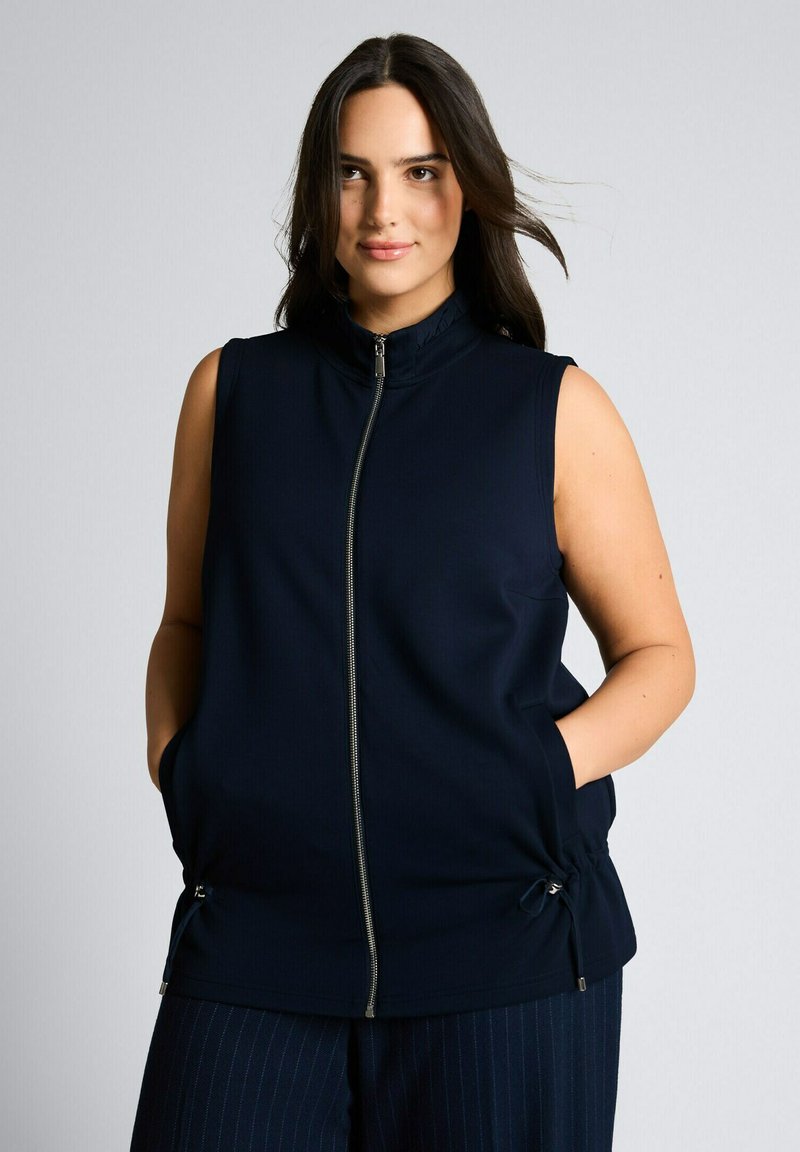 Hosszú sötét hajú nő, kék ujjak nélküli cipzáros mellényt visel, kezét zsebében tartja, világosszürke egyszínű háttér előtt áll.