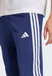 Σκούρο μπλε αθλητικό παντελόνι με λευκό σχεδιασμό τριών ριγών, ομαλή υφή και τσέπη με φερμουάρ, με λευκό λογότυπο Adidas στο μηρό.