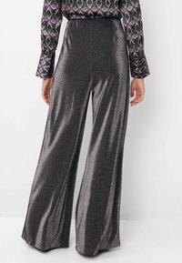 Vrouw in hoge taille, wijde metallic zilveren broek, gecombineerd met een langemouwen blouse met patroon, gezien van achteren.