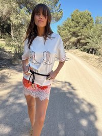 T-shirt branco com gráfico de botas de cowboy, saia curta vermelha com acabamento em renda branca e um cinto preto com detalhes em fivela de metal. Cenário natural ao ar livre.