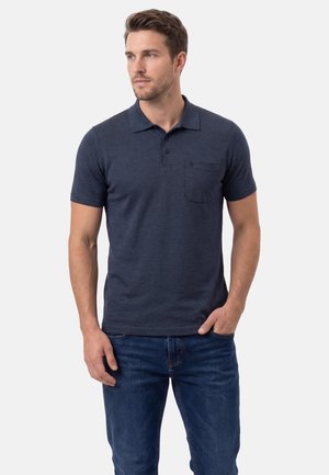 Mann trägt ein dunkelblaues Kurzarm-Poloshirt mit einer Brusttasche und blaue Jeans, steht mit einer Hand in der Tasche und schaut nach links.