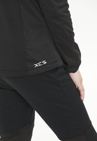 Sort sportsjakke og leggings med en glat tekstur, tætsiddende design og logoet "XCS" i hvid på jakens kant.