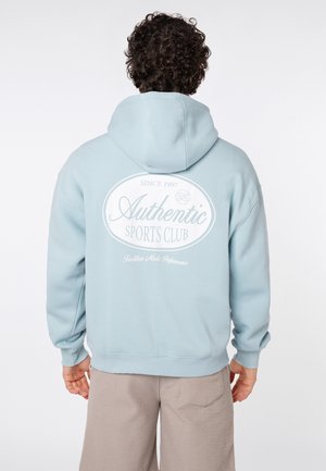 Person mit lockigem Haar, die einen hellblauen Hoodie mit dem Schriftzug „Authentic Sports Club“ und beigen Hosen trägt, vom Betrachter weggewandt.