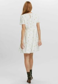 Robe blanche à manches courtes avec imprimé floral jaune, taille froncée, jupe évasée et détail d'ouverture en forme de clé au dos. Associée à des bottines noires.