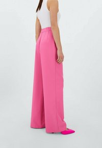 Kvinna iklädd högmidjade, vida rosa byxor, vit ärmlös topp och klarröda spetsiga klackskor, står i profil mot en neutral bakgrund.