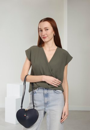 Olijfgroene wikkeltop met korte mouwen, gecombineerd met lichtblauwe high-waisted jeans. Zwart leren handtas in de vorm van een hart met ritssluiting.