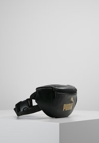 Černá kožená ledvinka s předním zipem, vyraženým zlatým logem, nastavitelným popruhem a kompaktním designem pro nošení nezbytností.