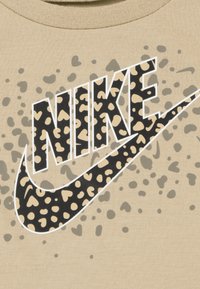 Tela beige con un estampado de leopardo negro y crema, con el logo de "Nike" que presenta patrones en forma de corazón y un contorno blanco. Diseño texturizado y casual.