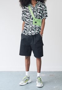 Person mit Afro-Haar trägt schwarze Shorts, ein grafisches Kurzarmhemd mit Augen- und Mundmustern, eine neongrüne Umhängetasche und Sneakers.
