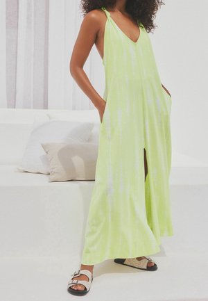 Femme portant une robe longue sans manches vert citron avec une fente avant et des poches, associée à des sandales beige et noir, debout à l'intérieur.