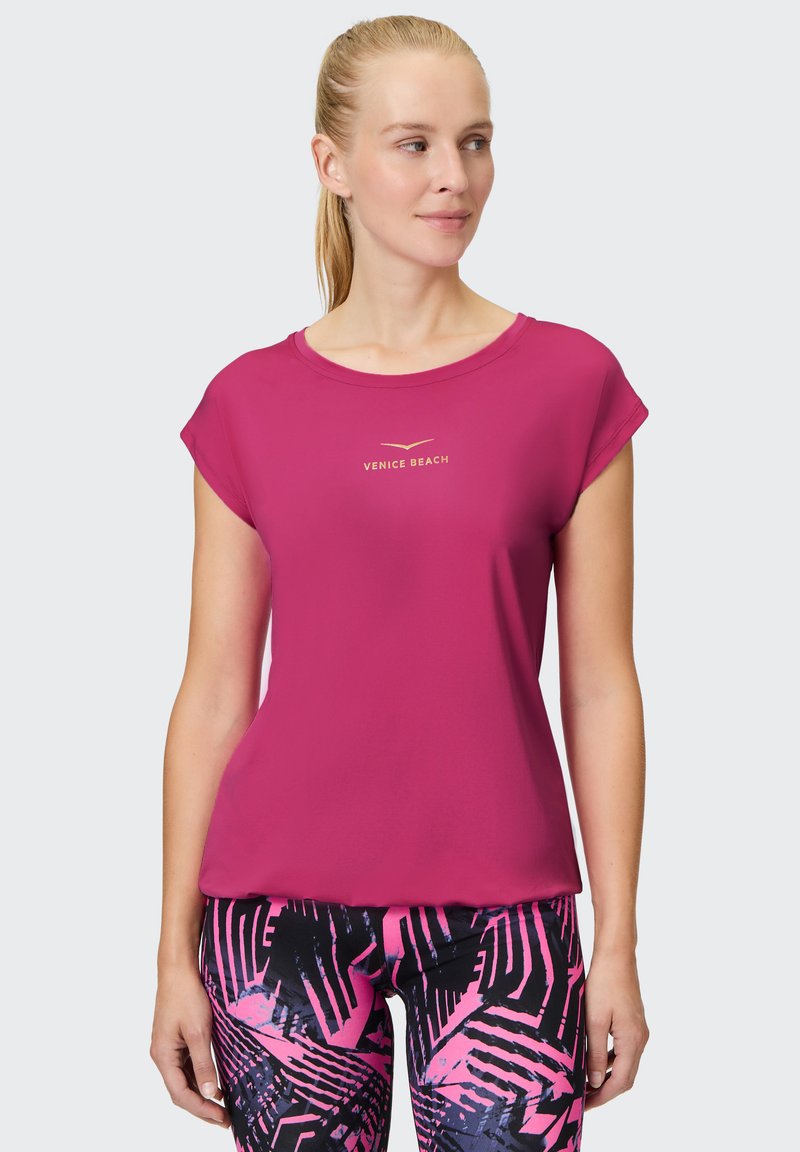 Fuchsienfarbenes Sport-T-Shirt mit kurzen Ärmeln, rundem Ausschnitt und "VENICE BEACH" Logo in Gold. Glatte Textur, kombiniert mit gemusterten Leggings.