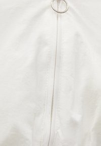 Biała kurtka o gładkiej fakturze, z centralnym zamkiem błyskawicznym z srebrną pętelką. Prosty design i minimalne detale.