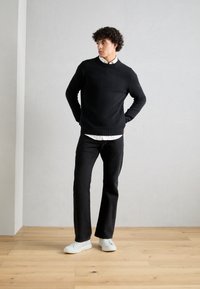Black knit sweater with a round neckline, paired with a white collared shirt, black straight-leg jeans, and white sneakers on a wooden floor.