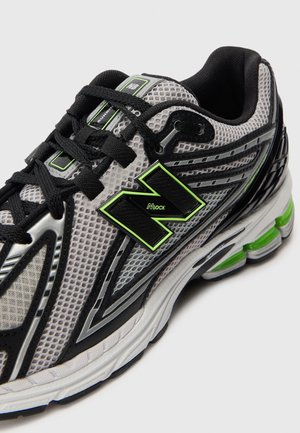 Personne portant un pantalon noir et des chaussures de course New Balance blanches avec des détails noirs et verts, debout sur un tapis bleu.