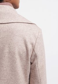 Persona vestida con un abrigo de lana beige, vista desde la parte posterior y lateral izquierdo, sobre un fondo blanco plaino.