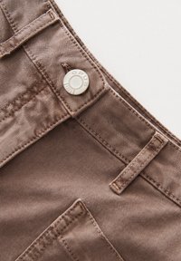 Bruin katoenstof met gedetailleerde stiksels, een metalen knop met "OPUS" gravure, en een platte tailleband met kleine lussen.