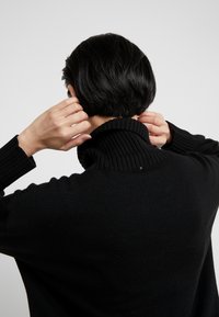 Personne aux cheveux courts noirs ajustant le col d'un pull à col roulé côtelé noir, vue de dos sur un fond uni.