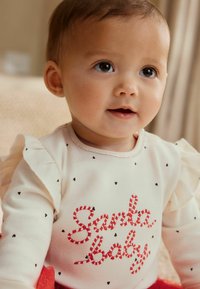 Cremefarvet langærmet skjorte med flæser på ærmerne, prydet med rødstribede "Santa baby"-tekster og sorte hjerte mønstre. Glat tekstur.