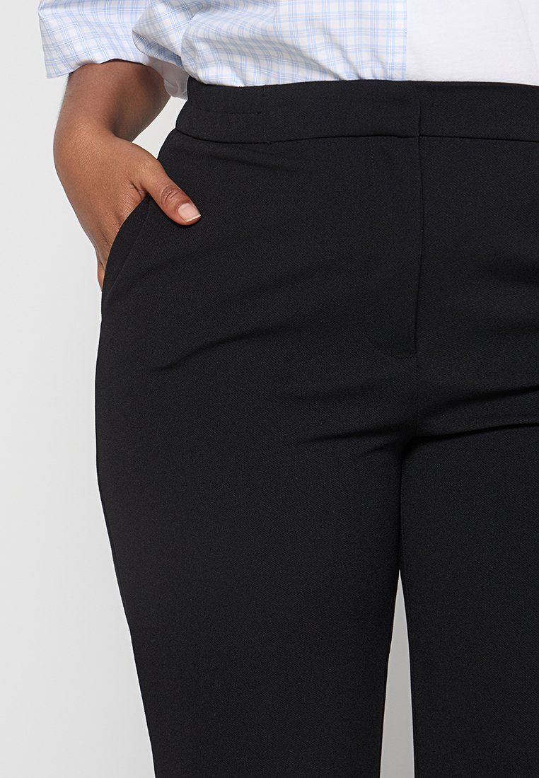 Person, der schwarze Hosen und ein in die Hose gestecktes hellblau kariertes Hemd trägt, mit einer Hand in der Tasche.