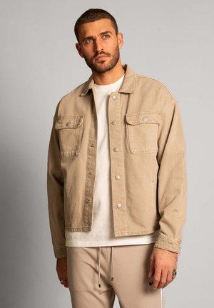 Beige denim jacket with button closures, two chest pockets, and a relaxed fit, layered over a white shirt and paired with beige pants.