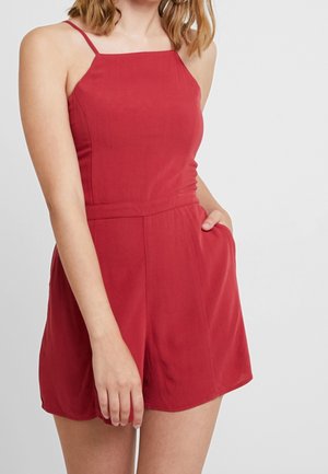 Frau trägt einen ärmellosen roten Overall mit dünnen Schulterriemen und Seitentaschen, steht vor einem einfarbigen hellen Hintergrund.