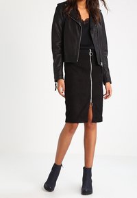 Veste en cuir noir avec des épaules texturées, haut en dentelle noire et jupe en daim noir longueur genou avec une fermeture éclair avant et un détail en anneau. Bottines noires.