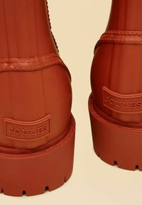 Gli stivali di gomma rossa presentano scanalature verticali, una suola texturizzata e un logo del marchio visibile sul retro. Hanno un design con plateau spesso.