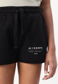 Pantalones cortos negros con cordón con las palabras "ALL SAINTS" y texto en japonés impresos sobre "LONDON" en la parte inferior del muslo izquierdo, usados por una persona con una camiseta de color claro.