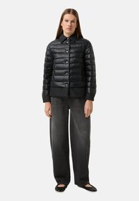 Schwarze Steppjacke mit glänzendem, gestepptem Finish, mit Stehkragen, Druckknöpfen und Seitentaschen, getragen über locker sitzenden Jeans.