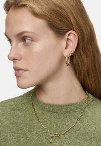 Goldfarbener Schmuck mit unregelmäßigen Formen: Creolen und eine strukturierte Kettenhalskette. Getragen mit einem grünen Strickpullover.