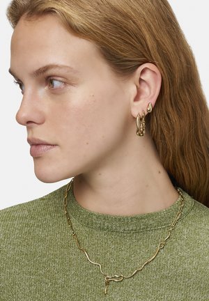 Goldfarbener Schmuck mit unregelmäßigen Formen: Creolen und eine strukturierte Kettenhalskette. Getragen mit einem grünen Strickpullover.