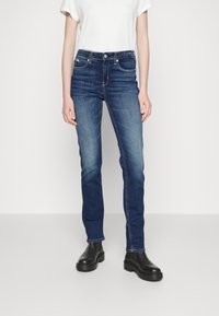 En person klädd i en vit kortärmad topp, blå slim-fit jeans och svarta ankelskorts stövlar, står mot en enkel vit bakgrund.
