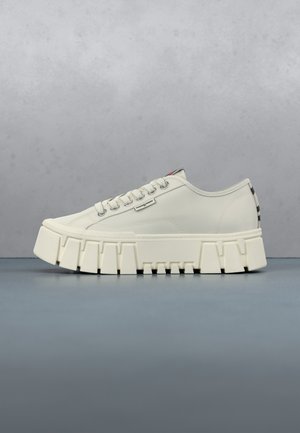 Witte platformsneaker met dikke gesegmenteerde zool, veters aan de voorkant en klein merketiket aan de zijkant, getoond op een grijze ondergrond en achtergrond.