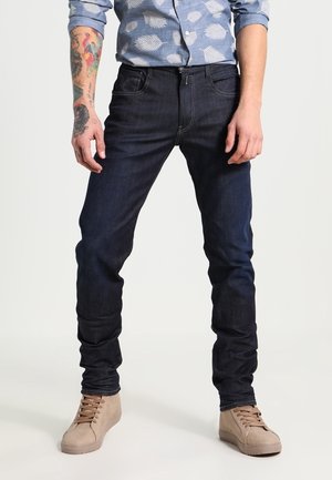 Vaqueros de color azul oscuro con un diseño ajustado, material de algodón y costuras mínimas. Combinados con zapatillas de deporte beige claras y una camisa azul con estampado.