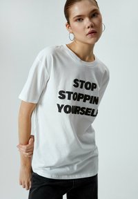 Hvid bomulds T-shirt med korte ærmer, der har fed sort tekst, som siger "STOP STOPPIN YOURSELF." Enkel rund halsudskæring og løs pasform.