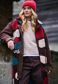 Žena opierajúca sa o kamenú stenu, oblečená v farebnej suchanej fleecovej bunde, béžovom pletenom svetri, tmavých nohaviciach a červenej čiapke.