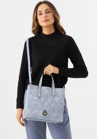Denim-stijl tote bag in lichtblauw met een textuur oppervlak, dubbele handgrepen, een afneembare schouderband en een cirkelvormige metalen accent aan de voorkant.